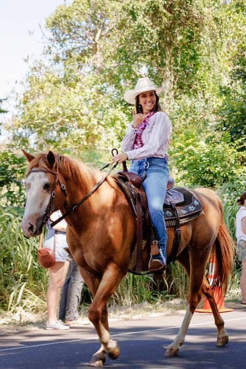 A woman riding a chestnut horse & wearing western attire wave during the 2025 Koloa Plantation Days Parade